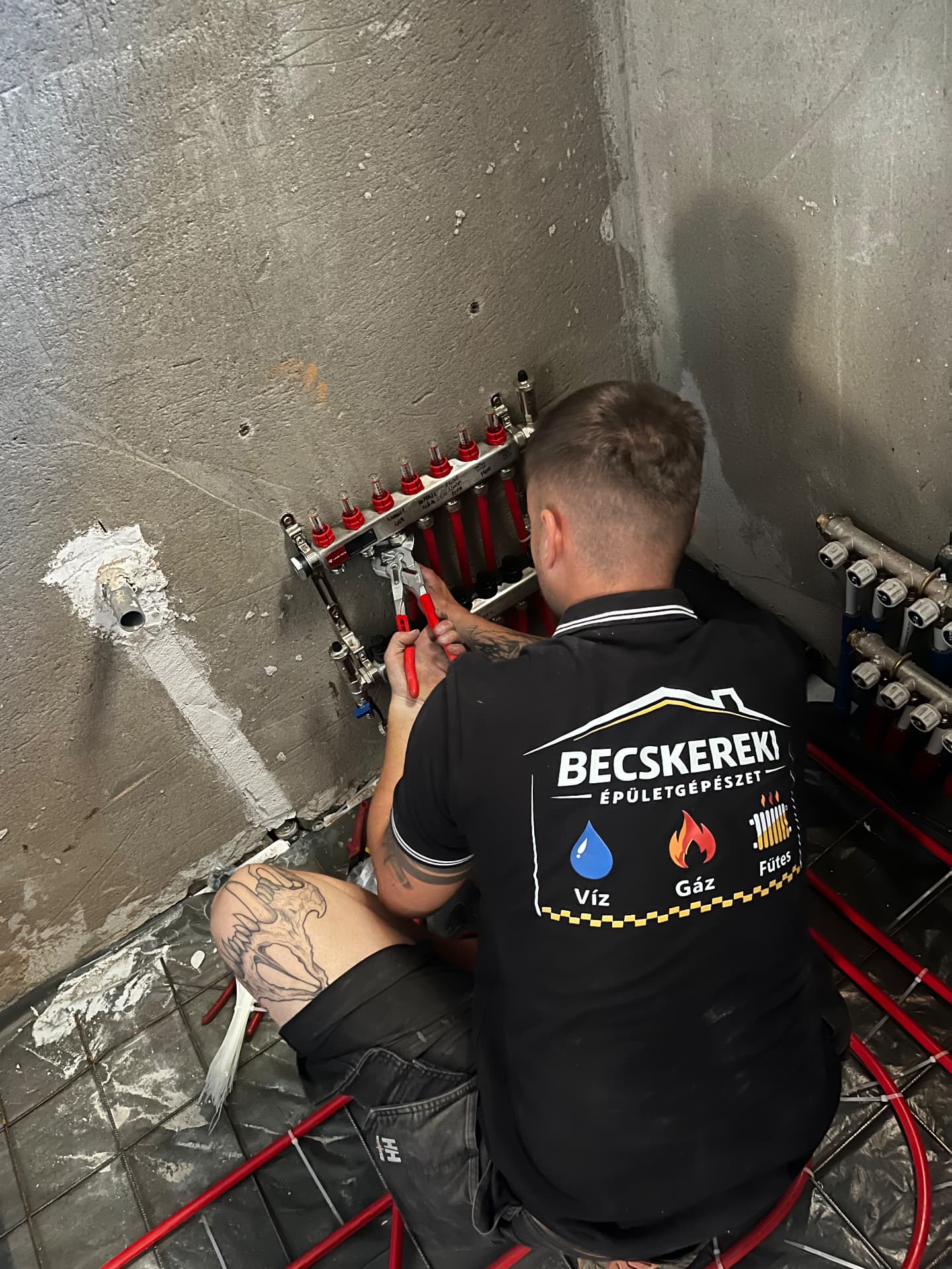 Szerelő padlófűtés-osztó-gyűjtőt szerel, a pólóján a Becskereki Épületgépészet felirat